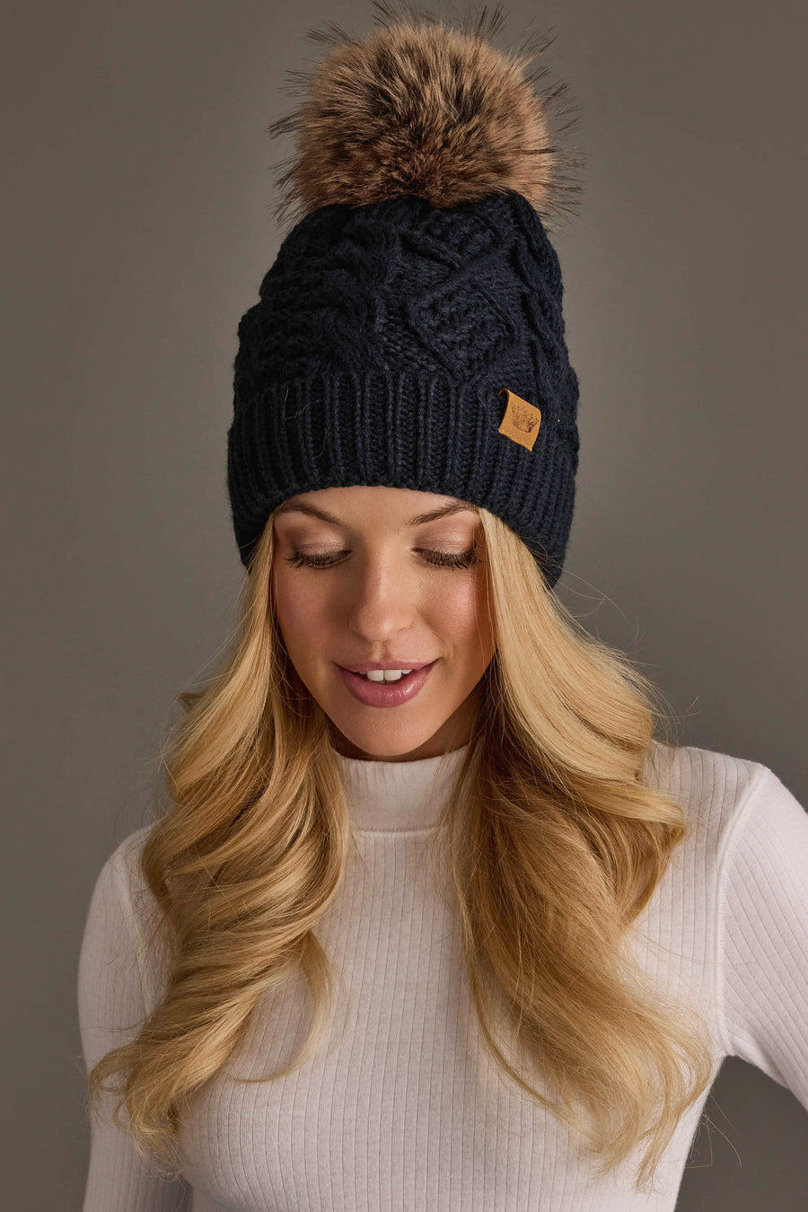 A woman with long blonde hair wears the Panache Apparel Collection Navy Cable Knit Pom Hat, a dark knit beanie topped with a large faux fur pom-pom, paired with a white ribbed turtleneck, as she looks down and smiles softly against a plain background.