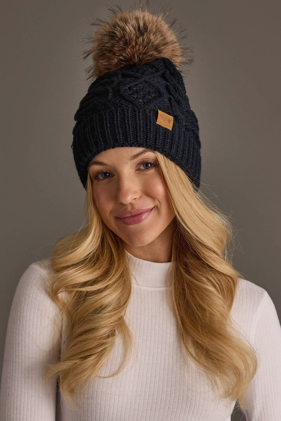 A woman with long blonde hair smiles softly against a plain gray background, wearing a white ribbed turtleneck and the Panache Apparel Collection’s Navy Cable Knit Pom Hat, which features a brown faux fur pom-pom.