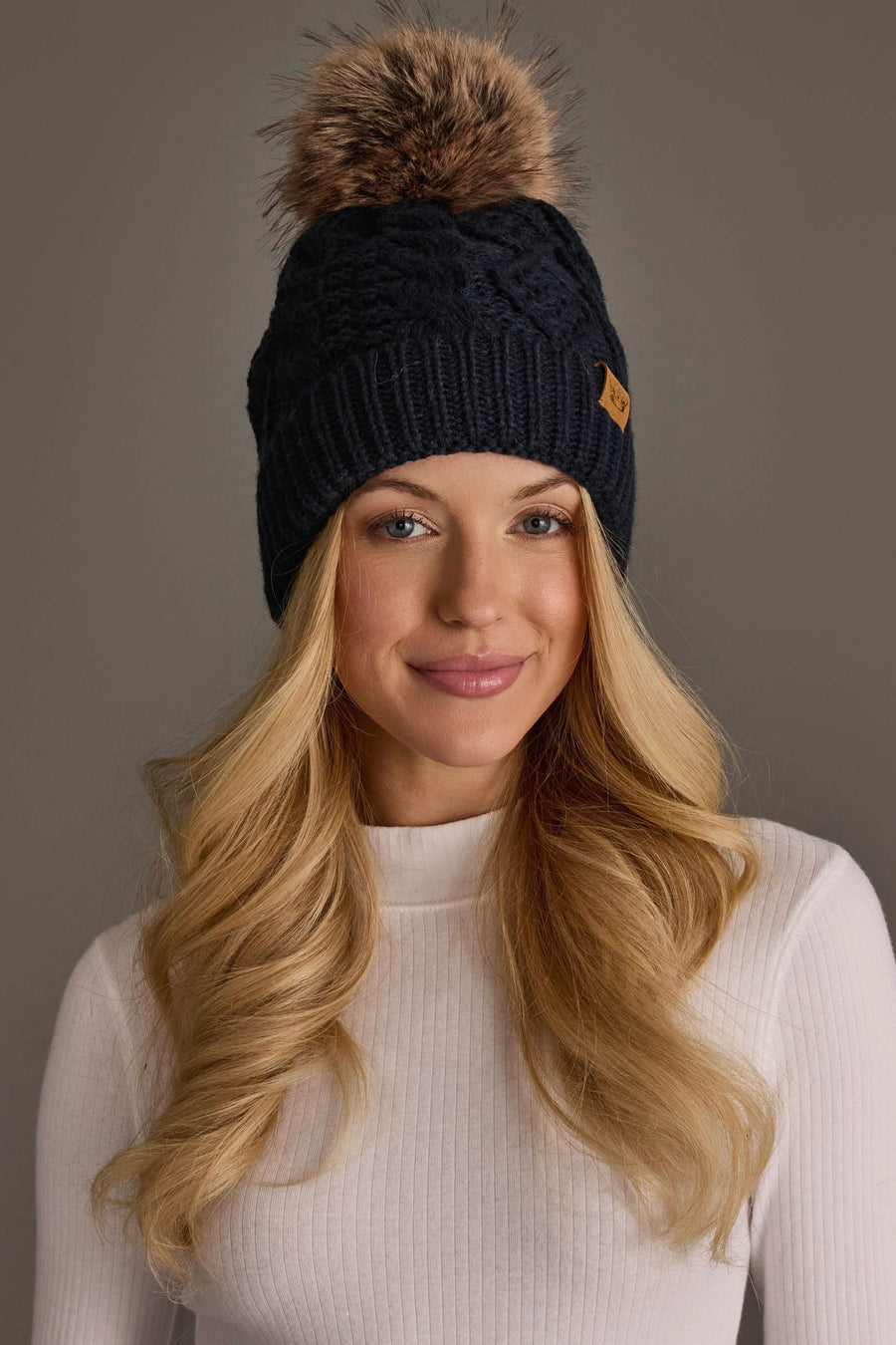 A woman with long blonde hair is smiling against a plain gray background while wearing a white ribbed sweater and the Panache Apparel Collection Navy Cable Knit Pom Hat, featuring a dark knit design topped with a faux fur pom-pom.