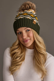 A woman with long blonde hair wears the Panache Apparel Collection Olive, Cream & Mustard Patterned Pom Hat, a fleece-lined knit beanie featuring a pom-pom and patterned design. She is dressed in a white ribbed top, smiling gently and looking off to the side against a plain background.