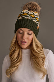 A woman with long blonde hair wears the Panache Apparel Collection’s Olive, Cream & Mustard Patterned Pom Hat, featuring a green knit design with a faux fur pom-pom and white patterning, paired with a white ribbed turtleneck sweater. She looks down with a neutral expression.