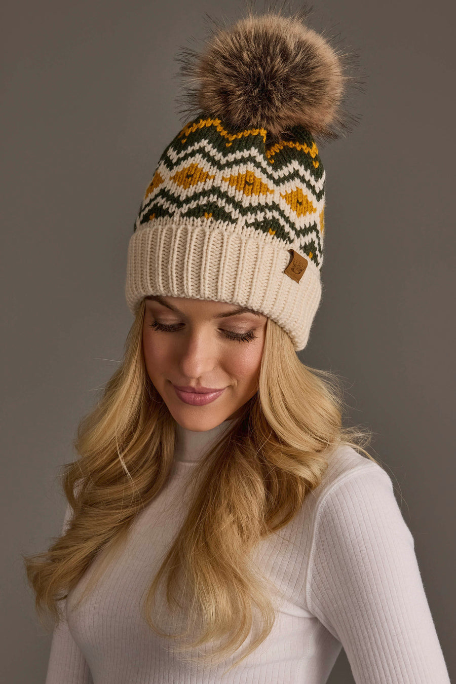A woman with long blonde hair wears a white ribbed top and the Cream, Olive & Mustard Patterned Pom Hat by Panache Apparel Collection, a fleece-lined knit hat adorned with a green, yellow, and white zigzag pattern and finished with a pom-pom, set against a plain grey background.