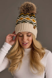 A woman with long blonde hair wears the Panache Apparel Collection Cream, Olive & Mustard Patterned Pom Hat, a cozy fleece-lined knit beanie with a bold yellow and green pattern. She is dressed in a white ribbed turtleneck, gently touching the side of the hat and smiling softly against a neutral background.