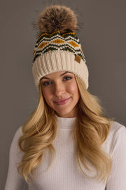 A woman with long blonde hair, dressed in a white sweater and the Panache Apparel Collection’s Cream, Olive & Mustard Patterned Pom Hat, a fleece lined knit winter hat featuring a faux fur pom-pom, smiles softly against a plain background.