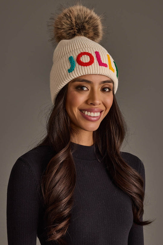 A woman wears a Beige Jolly Pom Hat, a beige knit hat with a faux-fur topper and the word JOLLY embroidered in multicolored letters.