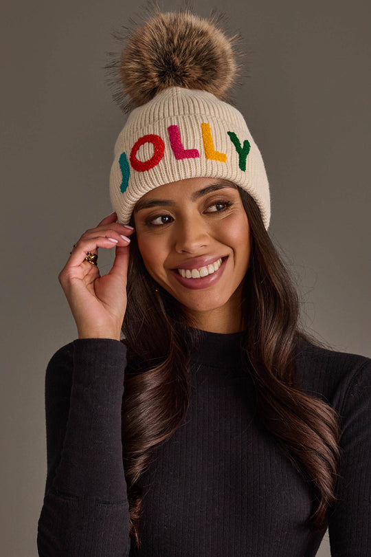 A woman wears a Panache Apparel Collection Beige Jolly Pom Hat, a beige knit hat with a faux fur pom-pom and the word JOLLY embroidered in multicolored letters.