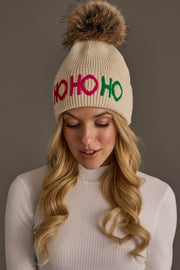 A Beige Ho Ho Ho Pom Hat featuring a beige knit design, a brown faux-fur pom-pom, and colorful HO HO HO embroidery.