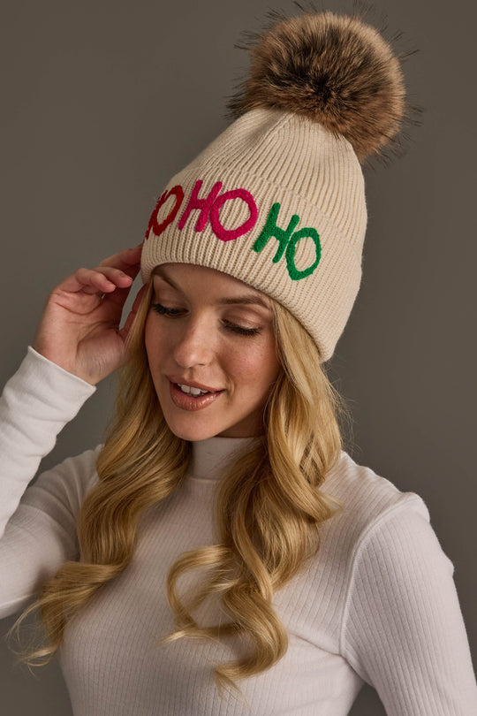 A person wearing a Beige Ho Ho Ho Pom Hat, which is a beige knit hat with a faux fur topper and the text HO HO HO in pink and green letters.