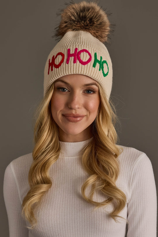 A person wearing a Beige Ho Ho Ho Pom Hat, featuring a beige knit design with a faux fur pom-pom and the text HO HO HO in colorful letters across the front.
