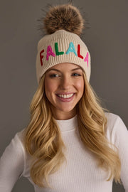 A person wearing a Beige Fa La La Pom Hat, a beige knit hat with a faux fur pom-pom and the word FALALA embroidered in colorful letters.