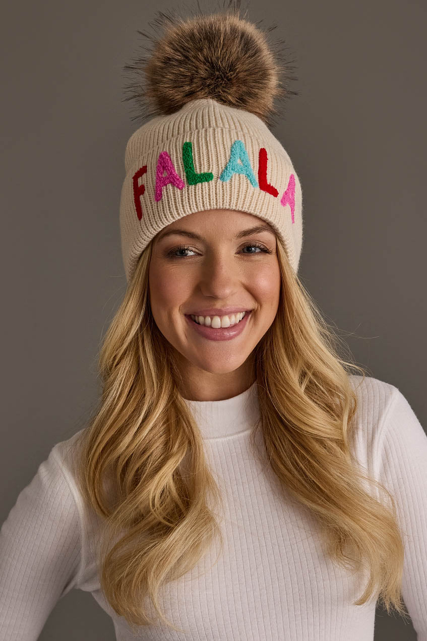 A person wearing a Beige Fa La La Pom Hat, a beige knit hat with a faux fur pom-pom and the word FALALA embroidered in colorful letters.