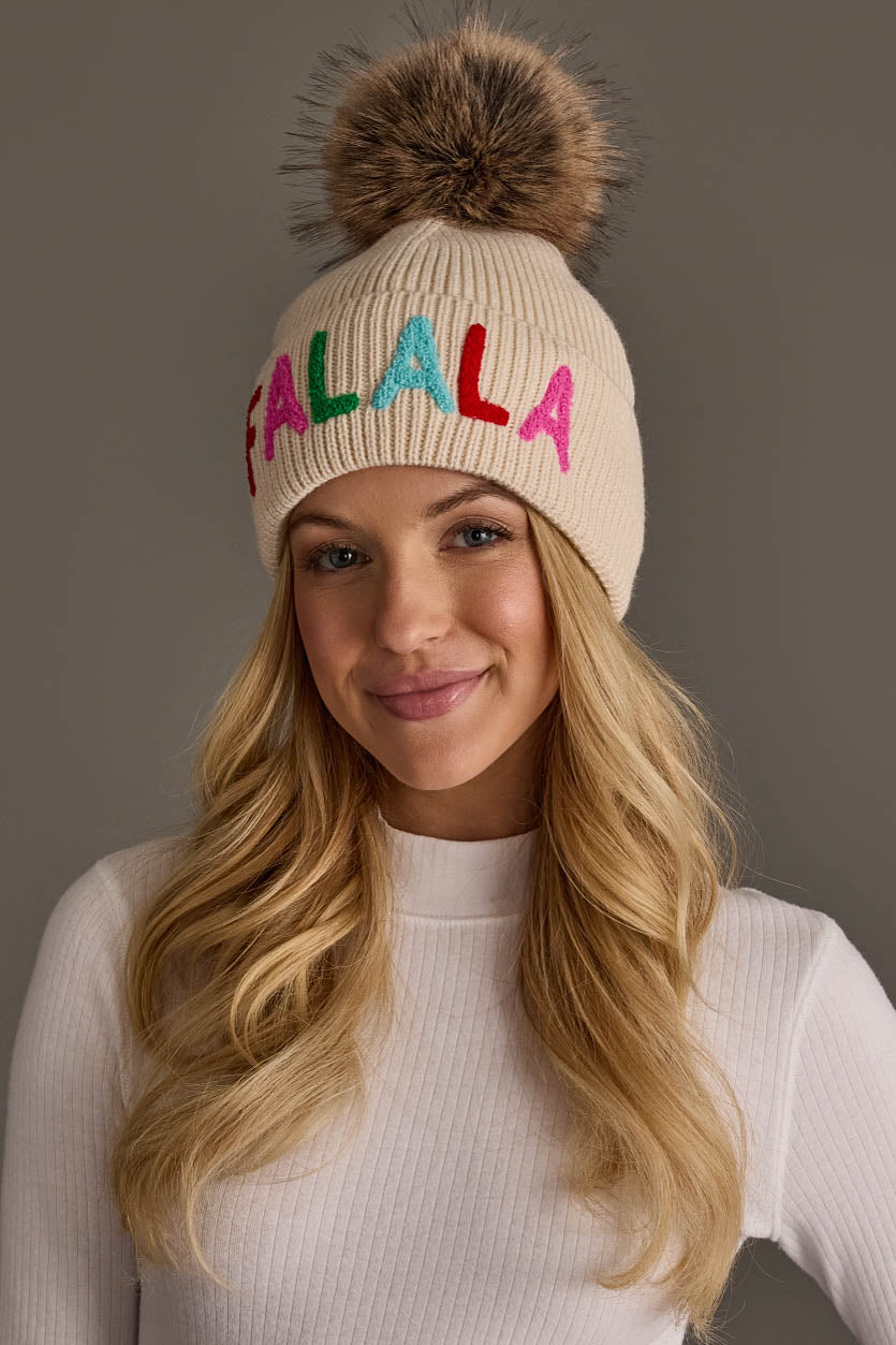 Beige Fa La La Pom Hat featuring a fluffy faux fur pom-pom and the text FA LA LA in colorful embroidered letters.