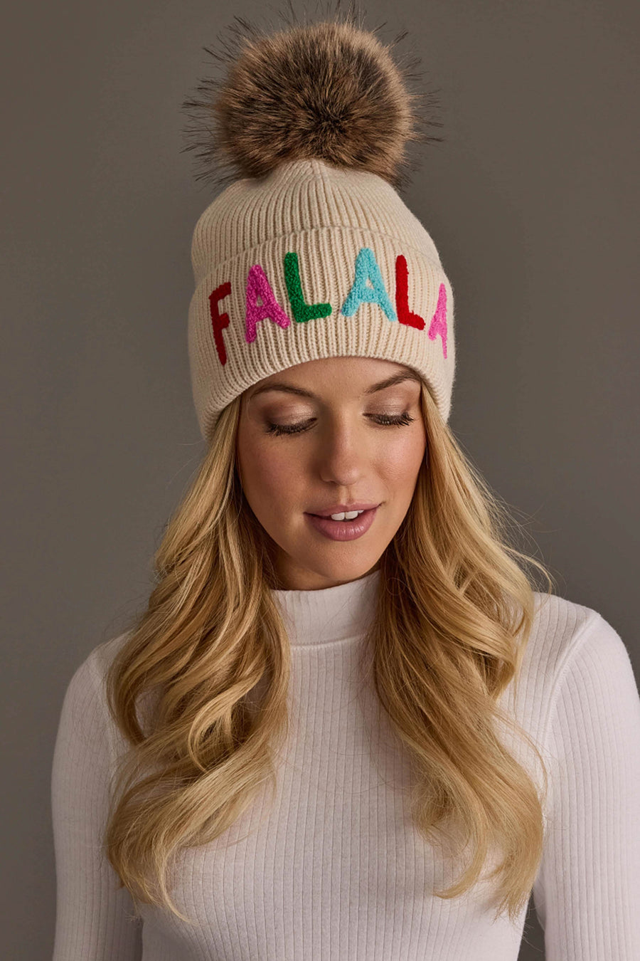 A person wearing a beige knit Beige Fa La La Pom Hat featuring a faux fur pom-pom and the word FALALA embroidered in colorful letters.