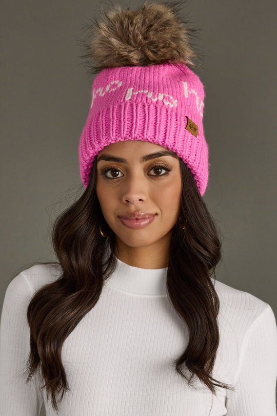 A woman with long wavy brown hair wears the Panache Apparel Collection Pink & White Ho Ho Ho Pom Hat, featuring a large faux fur pom-pom, white patterns, and cozy fleece lining, paired with a white ribbed turtleneck against a gray background.