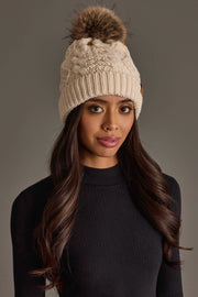 A woman with long, wavy brown hair wears the Ivory Cable Knit Pom Hat from Panache Apparel Collection, featuring a faux fur pom-pom, paired with a black ribbed long-sleeve top, standing against a neutral background.