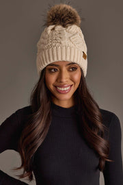A woman with long dark hair is smiling against a neutral background, wearing the Panache Apparel Collection Ivory Cable Knit Pom Hat, featuring a ribbed design and faux fur pom-pom, paired with a black long-sleeve top.