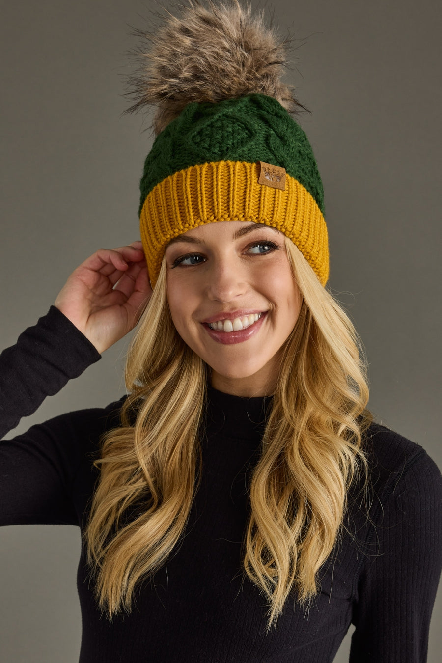 A smiling woman with long blonde hair wears the Panache Apparel Collection Green & Gold Trim Cable Knit Pom Hat, featuring a faux fur pom-pom. She is dressed in a black turtleneck and stands against a plain gray background.