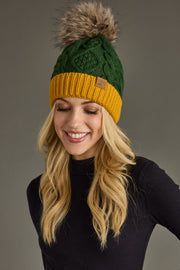 A smiling woman with long blonde hair wears the Panache Apparel Collection Green & Gold Trim Cable Knit Pom Hat, featuring a vibrant green knit design, mustard yellow brim, and a large faux fur pom, paired with a black turtleneck against a plain gray background.