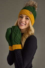 A woman smiles while wearing the Panache Apparel Collection Green & Gold Trim Cable Knit Pom Hat, featuring a faux fur pom-pom, along with coordinating green mittens with yellow cuffs and a black shirt, posing against a gray background.