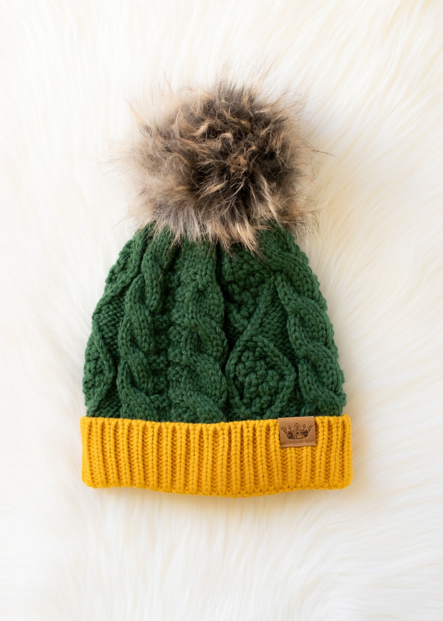 The Green & Gold Trim Cable Knit Pom Hat from Panache Apparel Collection features a green cable knit design with a gold ribbed brim and a brown faux fur pom-pom, fleece lined for extra warmth, displayed on a white fluffy surface.
