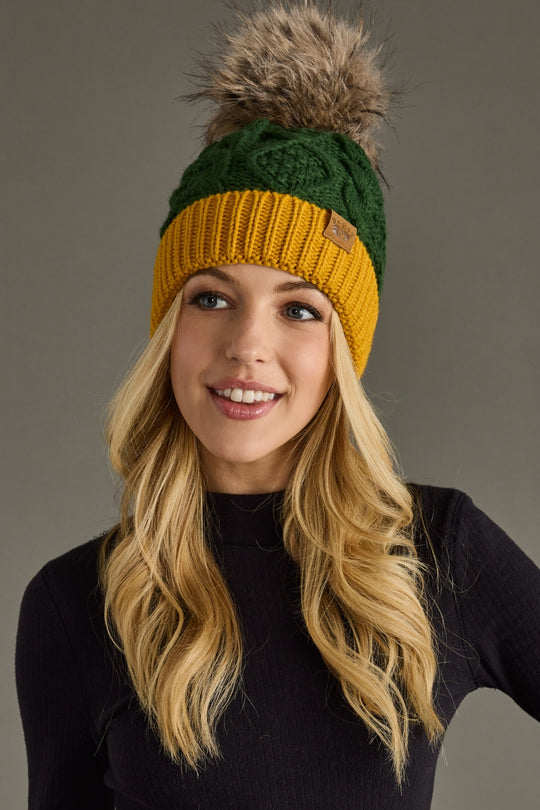 A smiling woman with long blonde hair wears the Panache Apparel Collection’s Green & Gold Trim Cable Knit Pom Hat, featuring a large faux fur pom-pom and cozy fleece-lined interior, paired with a black ribbed sweater. Gray background.