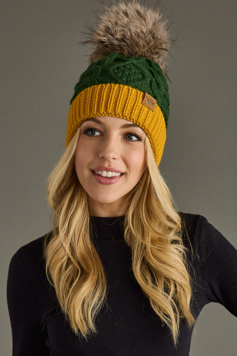 A smiling woman with long blonde hair wears the Panache Apparel Collection’s Green & Gold Trim Cable Knit Pom Hat, featuring a large faux fur pom-pom and cozy fleece-lined interior, paired with a black ribbed sweater. Gray background.