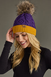 A woman in a black top smiles as she touches the Panache Apparel Collection Purple & Gold Trim Cable Knit Pom Hat, featuring a brown faux fur pom and cozy fleece lining, against a plain background.