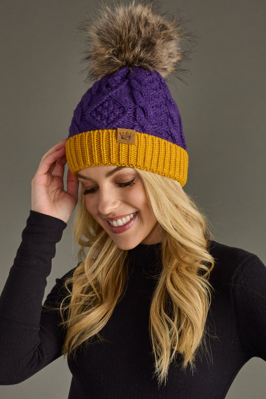 A woman in a black top smiles as she touches the Panache Apparel Collection Purple & Gold Trim Cable Knit Pom Hat, featuring a brown faux fur pom and cozy fleece lining, against a plain background.