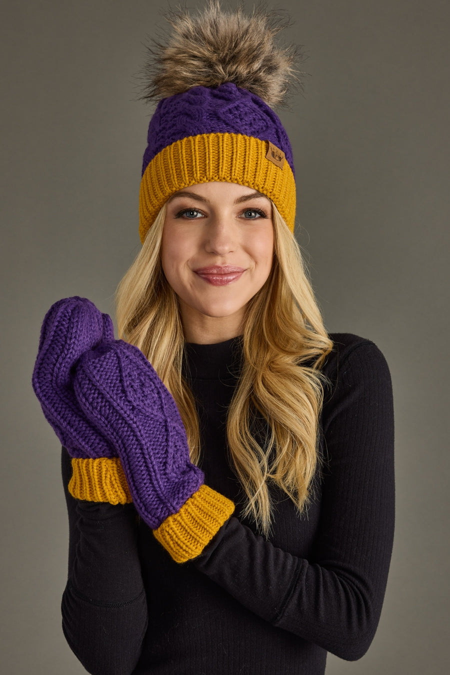 A smiling woman wears the Purple & Gold Trim Cable Knit Pom Hat by Panache Apparel Collection, featuring a fleece lining and faux fur pom. She pairs it with matching mittens, her long blonde hair visible beneath the hat, and a black sweater, while posing in front of a plain gray background.