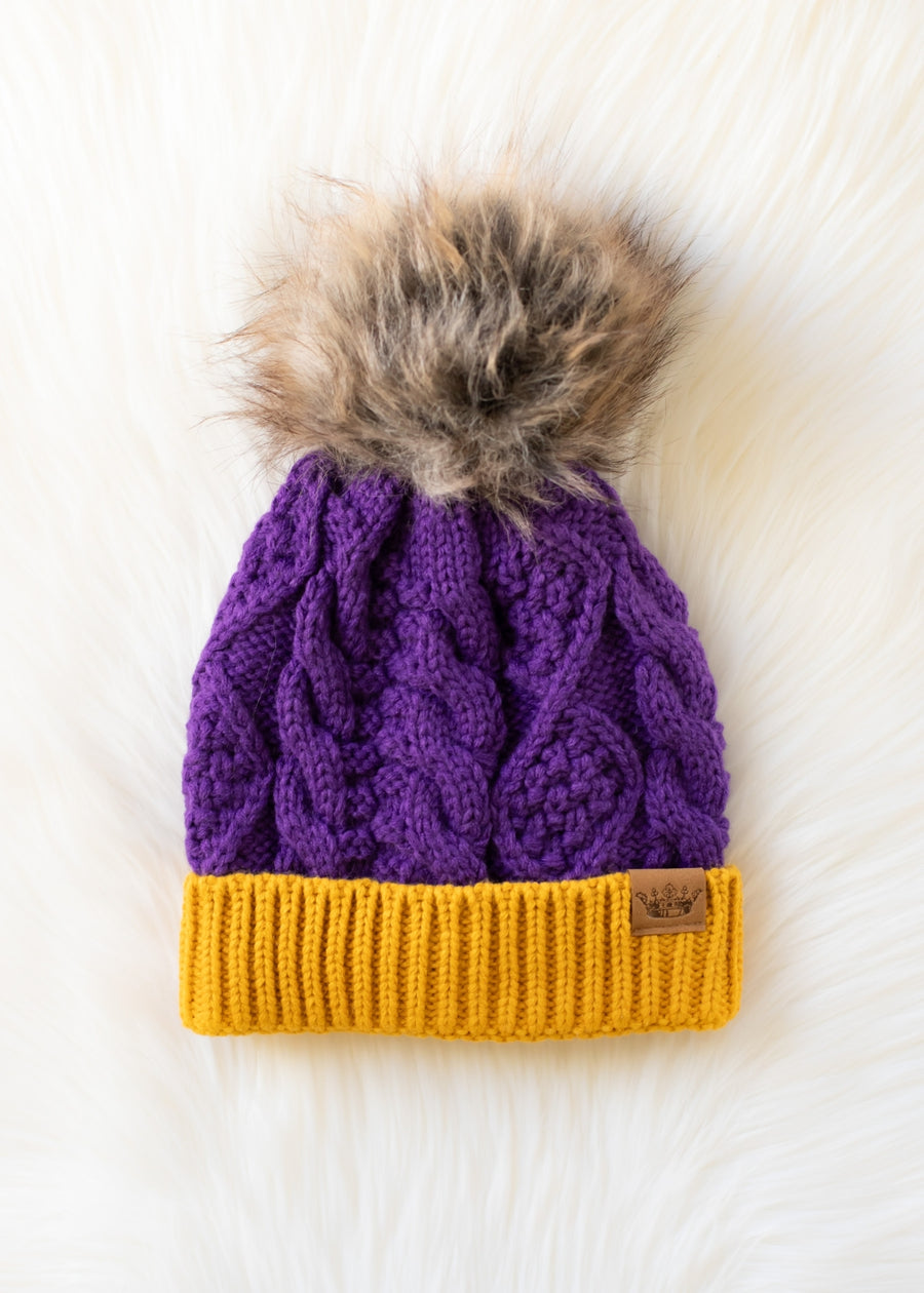 The Panache Apparel Collection Purple & Gold Trim Cable Knit Pom Hat, featuring a yellow ribbed brim and a large faux fur pom-pom on top, is fleece lined for extra warmth and shown resting on a soft white surface.