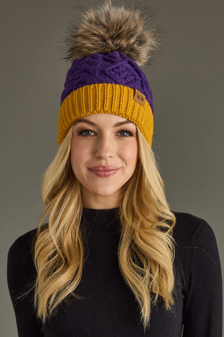 A woman with long blonde hair wears the Panache Apparel Collection Purple & Gold Trim Cable Knit Pom Hat, featuring a large faux fur pom, along with a black sweater. She smiles at the camera against a plain gray background.