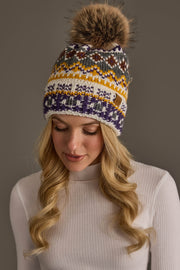 A woman with long blonde hair is wearing a white ribbed sweater and the Panache Apparel Collection Ivory, Purple & Gold Patterned Pom Hat, featuring a large faux fur pom; she looks down and smiles slightly.