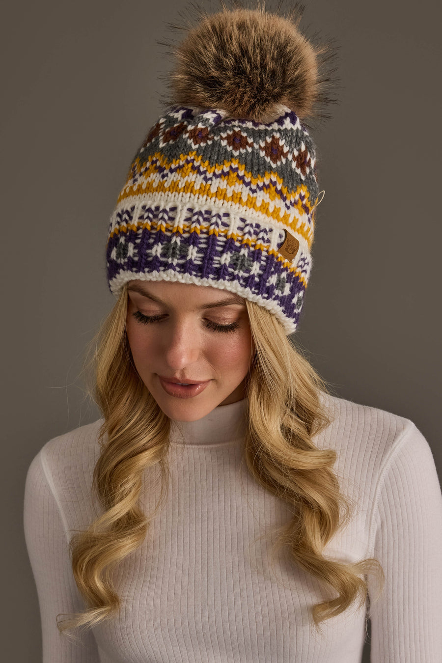 A woman with long blonde hair is wearing a white ribbed sweater and the Panache Apparel Collection Ivory, Purple & Gold Patterned Pom Hat, featuring a large faux fur pom; she looks down and smiles slightly.