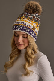 A woman with long blonde hair wears the Ivory, Purple & Gold Patterned Pom Hat from Panache Apparel Collection a colorful knit hat featuring a large faux fur pom along with a white ribbed turtleneck, looking down and smiling gently against a neutral background.