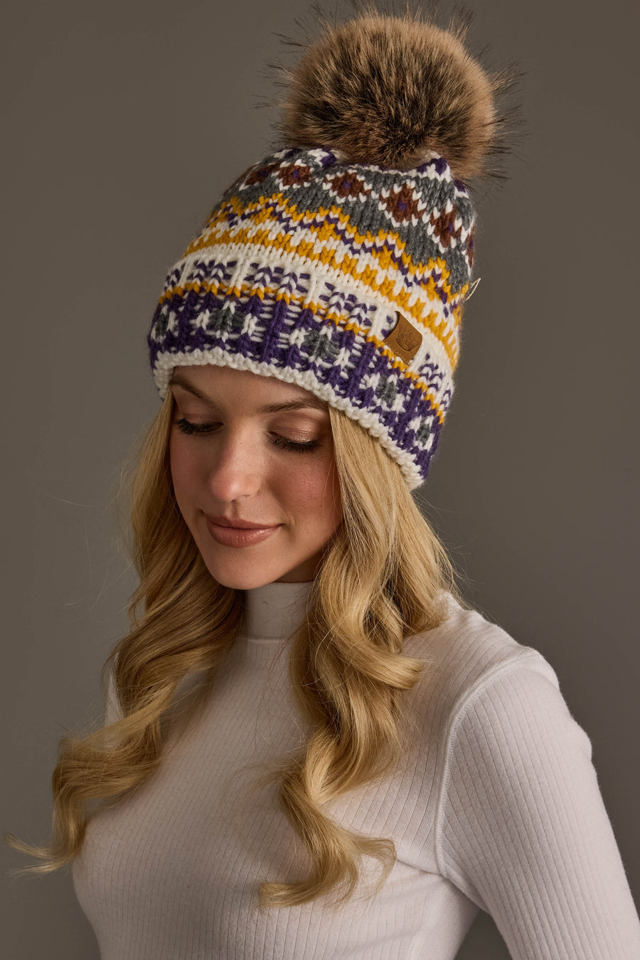 A woman with long blonde hair wears the Ivory, Purple & Gold Patterned Pom Hat from Panache Apparel Collection a colorful knit hat featuring a large faux fur pom along with a white ribbed turtleneck, looking down and smiling gently against a neutral background.