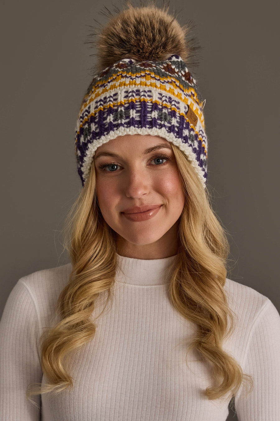A young woman with long blonde hair smiles softly against a plain, neutral background, wearing the Panache Apparel Collection's Ivory, Purple & Gold Patterned Pom Hat a colorful fleece-lined knit hat with a faux fur pom paired with a white ribbed turtleneck.