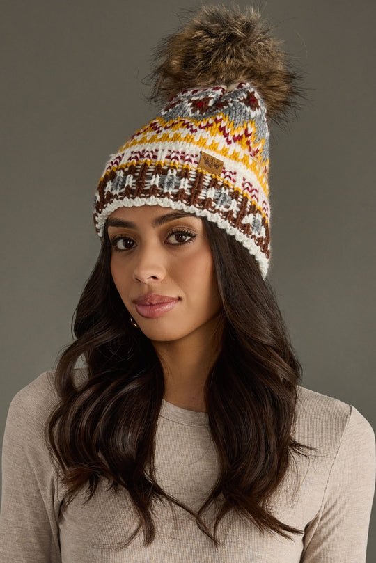 A woman with long dark hair, dressed in a beige shirt and wearing the Ivory, Red & Gold Patterned Pom Hat from Panache Apparel Collection a colorful, fleece-lined knit hat featuring a large faux fur pom poses against a plain gray background.