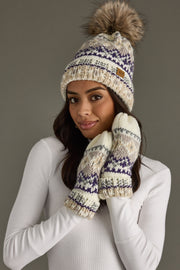 A woman in a white sweater wears the Panache Apparel Collection Ivory, Purple & Taupe Patterned Pom Hat with fleece lining and a faux fur pom, along with matching mittens, holding her hands near her face and smiling softly against a plain gray background.