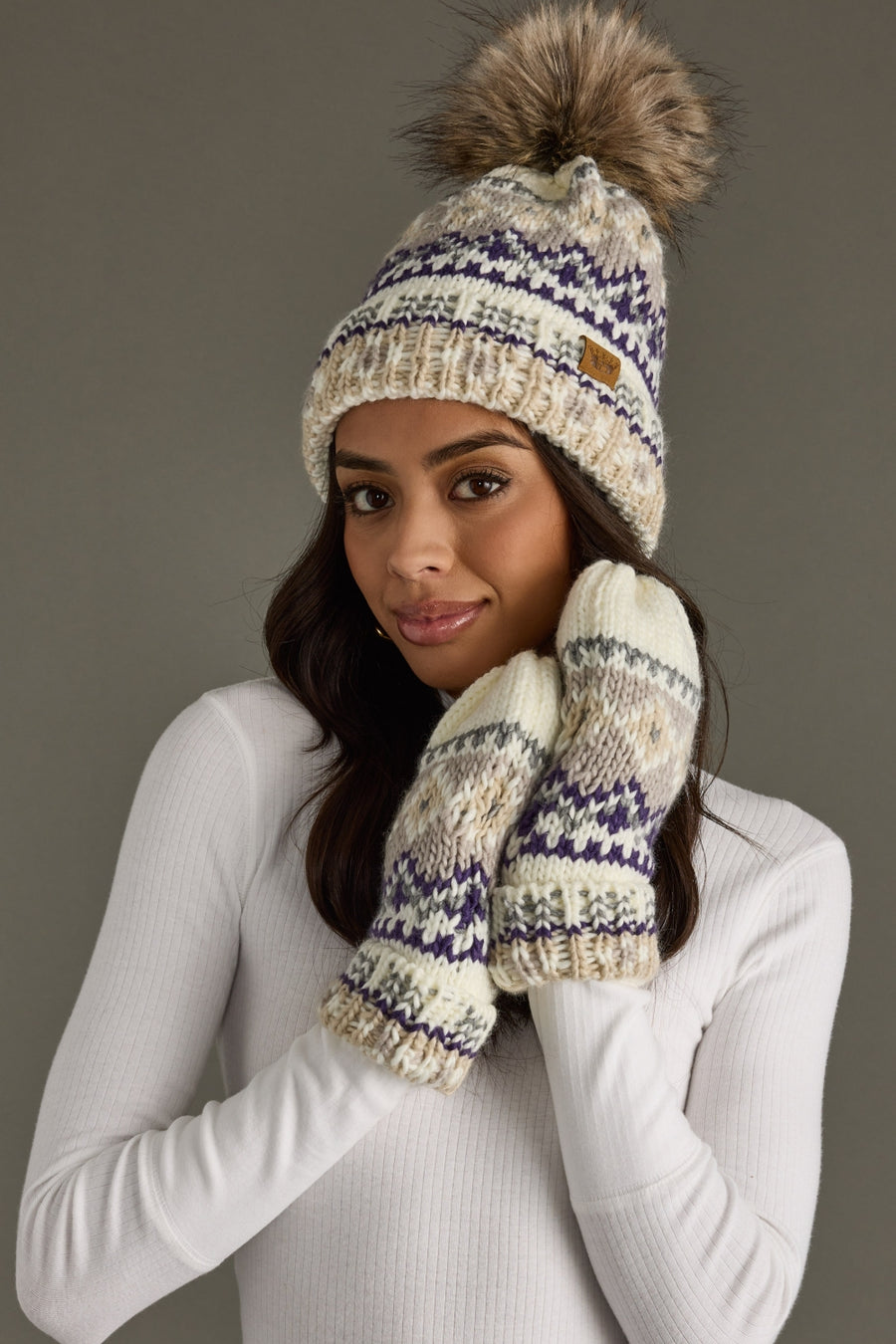 A woman in a white sweater wears the Panache Apparel Collection Ivory, Purple & Taupe Patterned Pom Hat with fleece lining and a faux fur pom, along with matching mittens, holding her hands near her face and smiling softly against a plain gray background.