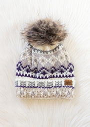 The Ivory, Purple & Taupe Patterned Pom Hat by Panache Apparel Collection features a cozy fleece lining, a stylish brown faux fur pom-pom, and an eye-catching cream, gray, and purple knit design. It's shown resting on a soft white surface.