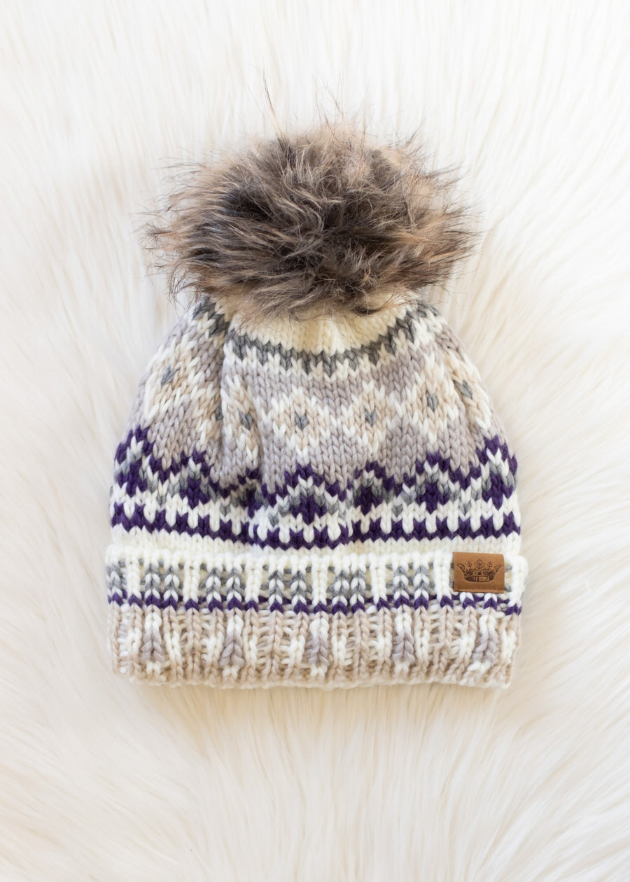 The Ivory, Purple & Taupe Patterned Pom Hat by Panache Apparel Collection features a cozy fleece lining, a stylish brown faux fur pom-pom, and an eye-catching cream, gray, and purple knit design. It's shown resting on a soft white surface.