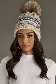 A woman with long dark hair wears the Panache Apparel Collection Ivory, Purple & Taupe Patterned Pom Hat—a cozy, fleece-lined knit hat featuring a large faux fur pom and purple accents. She pairs it with a white ribbed top and stands against a neutral gray background, looking at the camera.