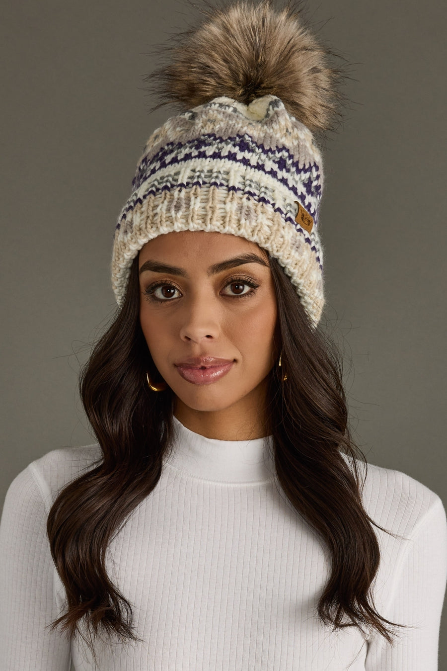 A woman with long dark hair wears the Panache Apparel Collection Ivory, Purple & Taupe Patterned Pom Hat—a cozy, fleece-lined knit hat featuring a large faux fur pom and purple accents. She pairs it with a white ribbed top and stands against a neutral gray background, looking at the camera.