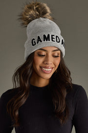 Woman modeling Panache Apparel’s gray Game Day knit beanie with black lettering and faux fur pom.