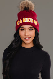 A woman with long black hair is wearing the Panache Apparel Collection Maroon & Gold Game Day Pom Hat, featuring a fur pom-pom and bold GAMEDAY text. She is dressed in a black top and standing against a plain gray background. This cozy, fleece-lined beanie ensures warmth and style wherever she goes.