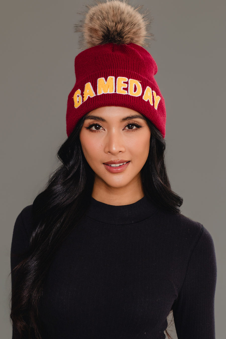 A woman with long black hair is wearing the Panache Apparel Collection Maroon & Gold Game Day Pom Hat, featuring a fur pom-pom and bold GAMEDAY text. She is dressed in a black top and standing against a plain gray background. This cozy, fleece-lined beanie ensures warmth and style wherever she goes.