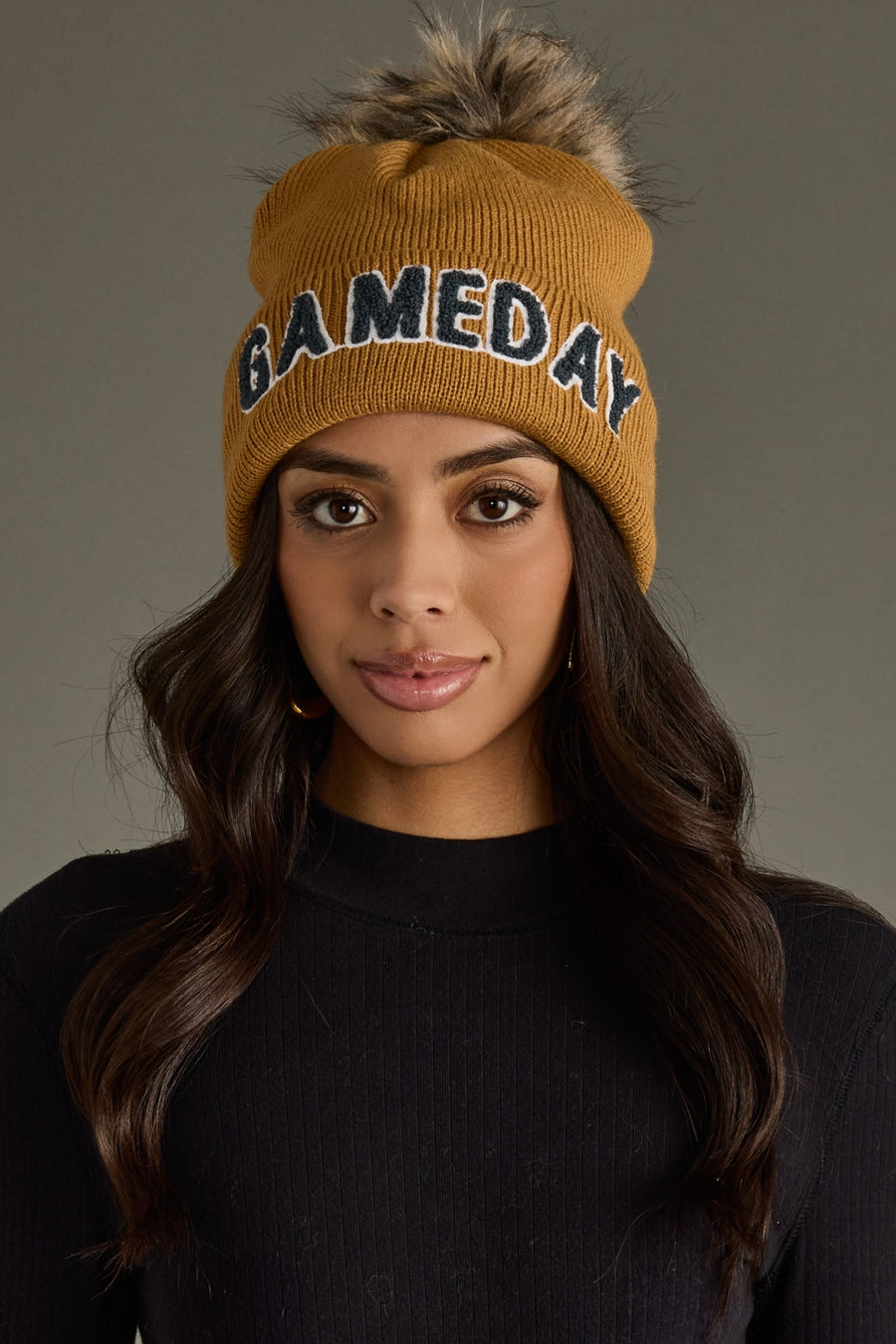 Woman with long dark hair wearing Panache Apparel’s camel and gray Game Day pom hat with bold lettering and a faux fur pom.