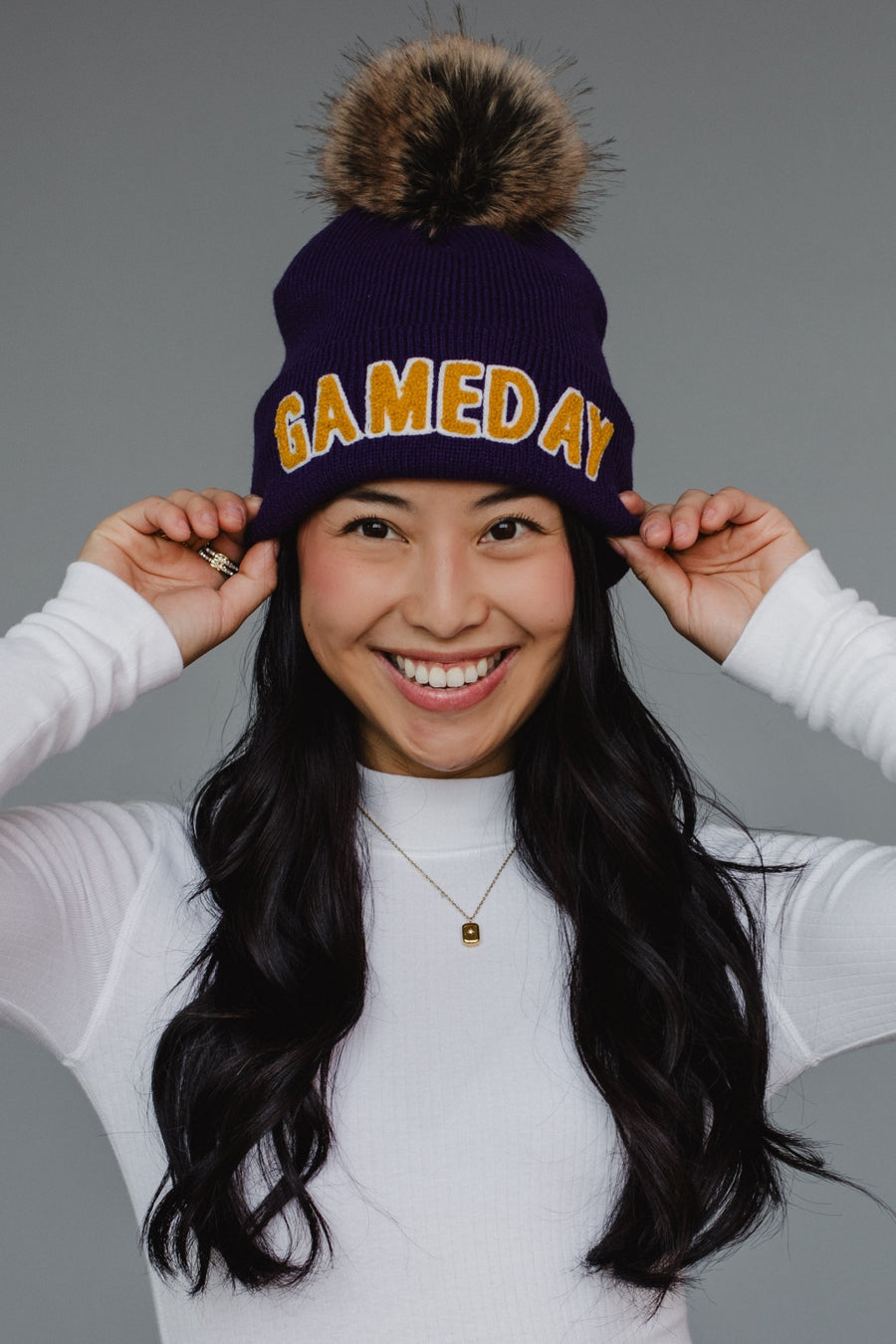 A navy blue ribbed knit beanie features bold "GAMEDAY" lettering in gold chenille across the cuff, topped with a luxurious faux-fur pom-pom in brown and black tones. The classic cuffed design combines spirited collegiate styling with cozy winter functionality, perfect for game day or everyday cold-weather wear.