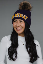 A navy blue ribbed knit beanie features bold "GAMEDAY" lettering in gold chenille across the cuff, topped with a luxurious faux-fur pom-pom in brown and black tones. The classic cuffed design combines spirited collegiate styling with cozy winter functionality, perfect for game day or everyday cold-weather wear.