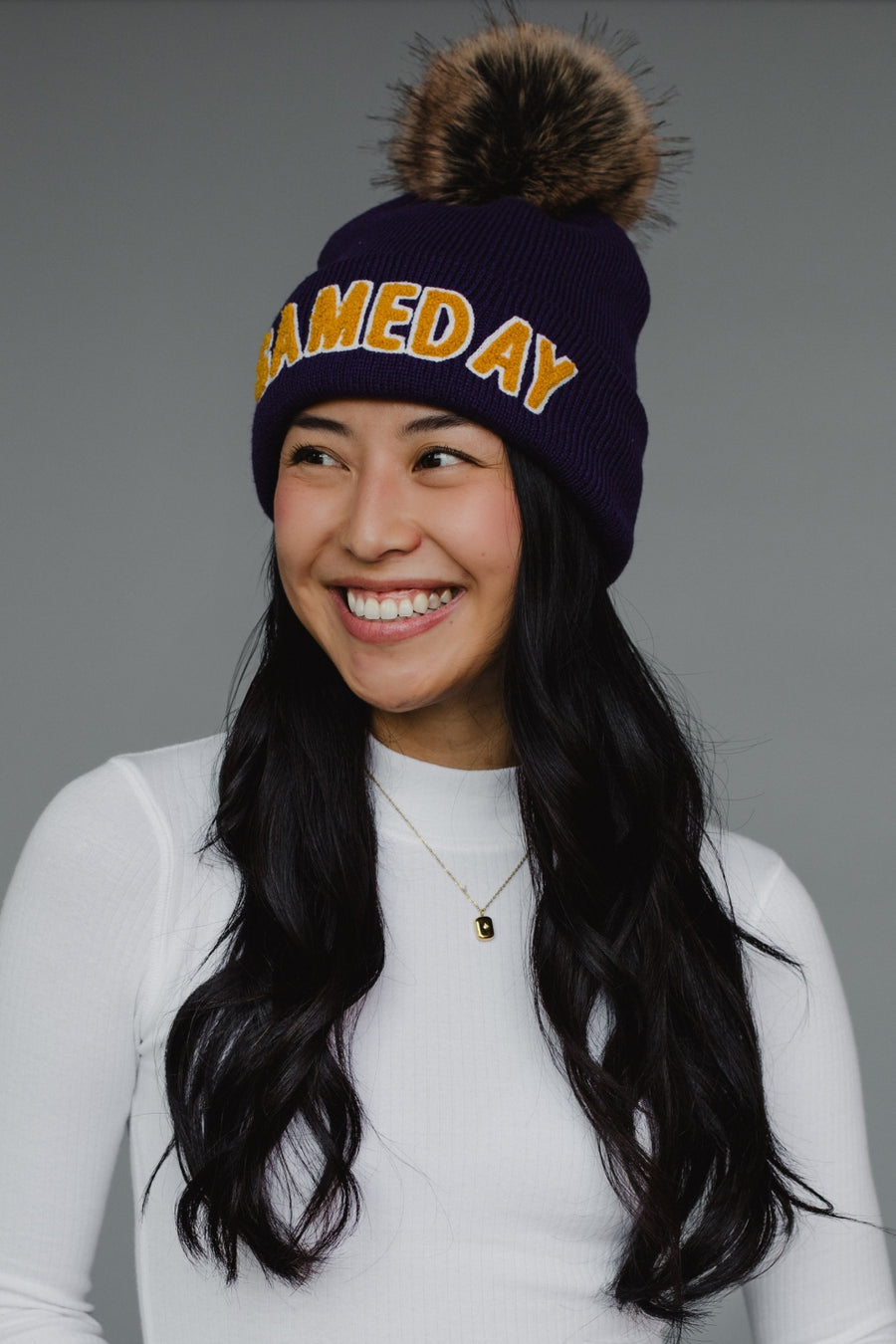 A navy blue ribbed knit beanie features bold "GAMEDAY" lettering in gold chenille across the cuff, topped with a luxurious faux-fur pom-pom in brown and black tones. The classic cuffed design combines spirited collegiate styling with cozy winter functionality, perfect for game day or everyday cold-weather wear.